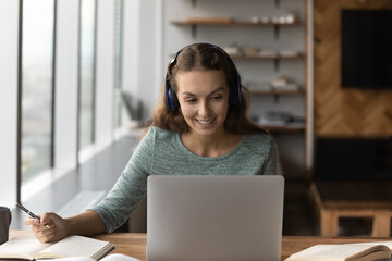 Happy young woman in headphones look at laptop screen make notes write in notebook consult client or customer online. Smiling female study distant on computer, talk on webcam video zoom call.