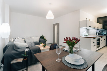 Living room connected with dining area and kitchen. Interior is decorated in gray. Room is dominated by wooden materials and modern workmanship. Plants predominate among the decorative elements.