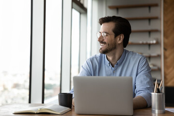 Overjoyed male employee sit at desk in office work on laptop look in distance thinking dreaming of success or opportunities. Happy young businessman plan or visualize. Business vision concept.