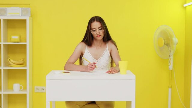 Young woman, manager, is sitting at work place and practices brainwriting, trying to find best solution of the issue, making notes on paper sheet, electric fan is working in the corner, foreground.