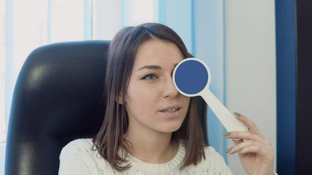 A young Caucasian woman in a cozy white sweater in the ophthalmologist's office at the optics clinic answers a doctor's question during a vision test.