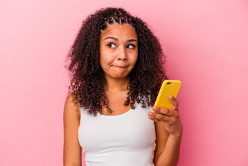 Young african american woman holding a mobile phone isolated a pink background confused, feels doubtful and unsure.