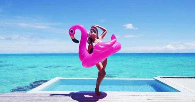 Summer suntan woman wearing hat and sunglasses for sun protection while sunbathing on Caribbean beach vacation by luxury infinity swimming pool. Fun flamingo pool toy air float