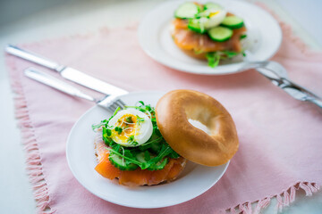 Fresh bagel sandwiches with salmon, soft egg, salad, microgreen sprouts, cucumber and cream cheese served on pink napkin with cutlery on the white kitchen table. Healthy Breakfast. Selective focus