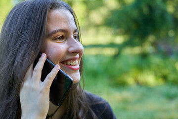 long-haired, fair-skinned woman sitting at a stone table with a coffee and her bag while talking on the phone, with green unfocused background. teenager in nature with his mobil taking a break.