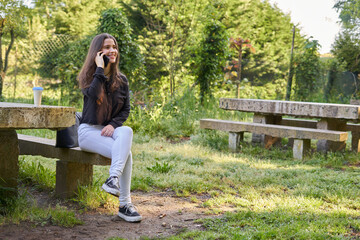 long-haired, fair-skinned woman sitting at a stone table with a coffee and her bag while talking on the phone, with green unfocused background. teenager in nature with his mobil taking a break.