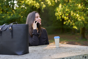 long-haired, fair-skinned woman sitting at a stone table with a coffee and her bag while talking on the phone, with green unfocused background. teenager in nature with his mobil taking a break.