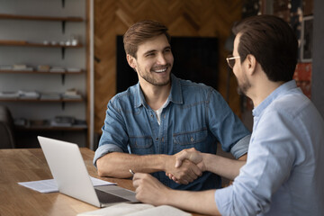 Smiling Caucasian young businessmen shake hands get acquainted greet at office meeting. Happy male...