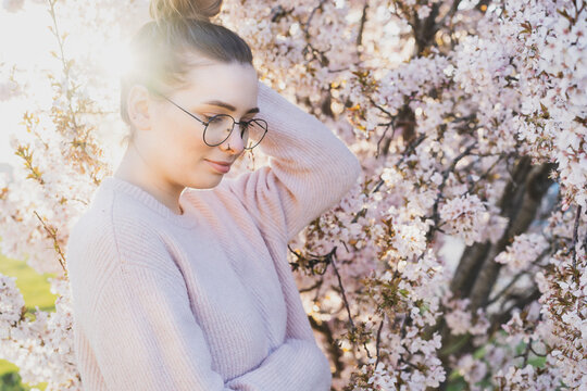 Portrait of young caucasian brunette hipster girl in glasses and light pink clothes near blooming tree with sunset light and sunbeams. Generation Z girl enjoy spring mood. Selective focus, copy space.