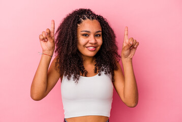 Young african american woman isolated on pink background indicates with both fore fingers up...