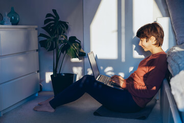 Mixed race woman sitting on the floor in her bedroom using a laptop in sunset light and shadows. Remote work at home. Woman scrolling social networks, chatting, watches video. Copy space.