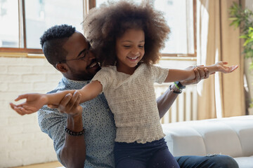 Happy African American dad teaching biracial preschool daughter to dance, holding girls flying hands and speaking. Daddy spending leisure time for kid training, development, active entertainment