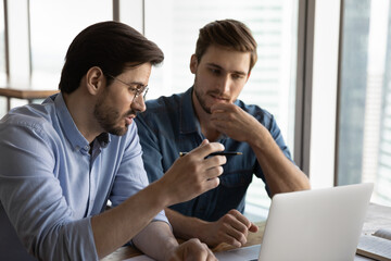 Concentrated young Caucasian businessmen work together on computer at workplace, engaged in team...
