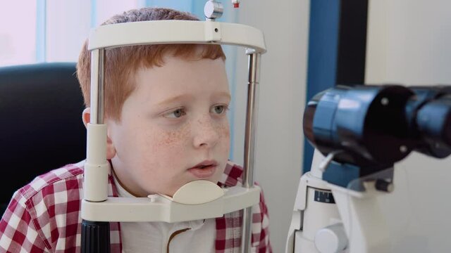 A red-haired caucasian boy with braces in a checkered red and white shirt in the ophthalmologist's office in the optics clinic undergoes a vision test using an autorefractometer