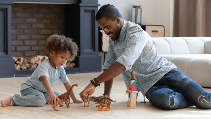Happy daddy and toddler son playing games with small dinosaurs together, enjoying weekend in comfortable cozy house, having fun. Little boy and hos dad spending leisure time, sitting on heat floor
