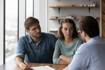 Unhappy young Caucasian couple buyers talk with male realtor or real estate agent dissatisfied with...