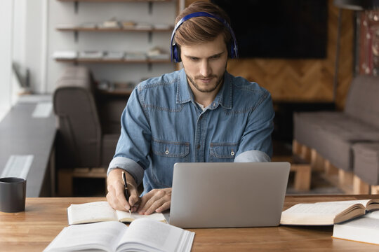 Serious Man In Earphones Busy Working On Laptop Consult Client Customer Online On Video Call. Male Employee Or Student Take Notes Write Study Learn On Computer, Have Webcam Zoom Conference.