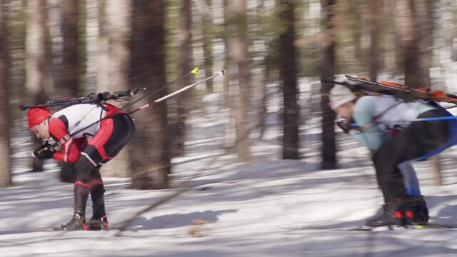 PAN of group of biathletes with poles and rifles skiing on the cross country track in forest while having workout