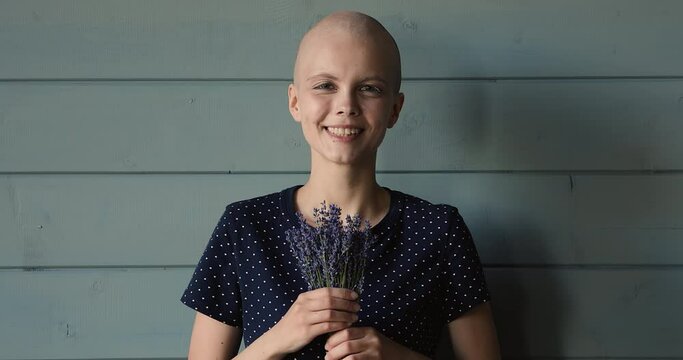 Happy young woman with bald after chemotherapy head holding bunch of lavender flowers, looking at camera, feeling hope for recovery or inspiration for cancer fight, isolated on studio background.