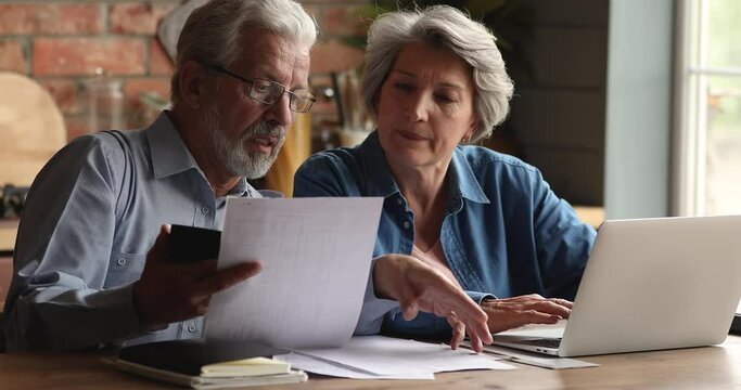 Concentrated mature older family couple using computer e-banking application, paying utility bills or taxes online, managing monthly household budget or calculating expenses together at home.