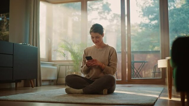 Smiling Young Woman Using Smartphone at Home, does Remote Work. Beautiful Girl Sitting on the Floor Uses Mobile Phone Internet, e-Shopping, Order Products Online, Post on Social Media. Sunny Home