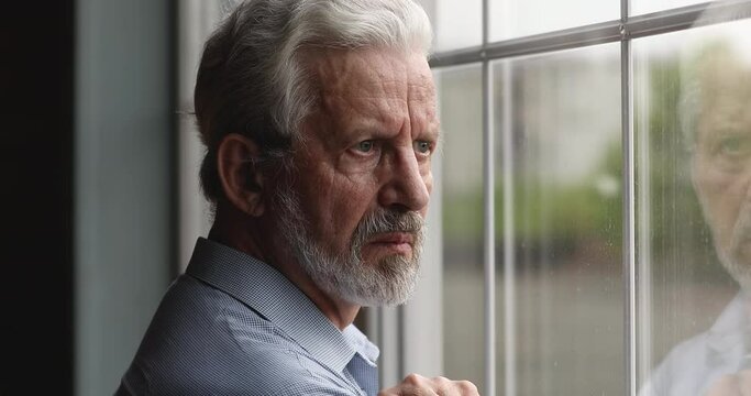 Frustrated middle aged 60s retired man taking off eyeglasses, looking in distance out of window, thinking of personal problems, suffering from loneliness or negative thoughts indoors, ageing concept.