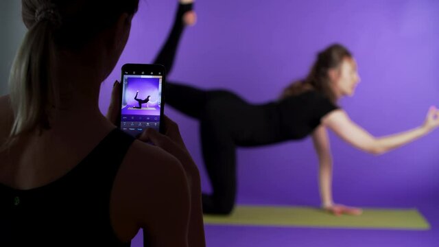 Woman is taking photos and making video of another woman who is posing on the yoga mat. Violet background