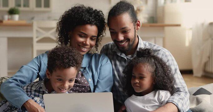 Happy bonding young mixed race couple parents teaching small adorable kids sibling using laptop software educational applications or enjoying leisure choosing cartoons, resting together at home.