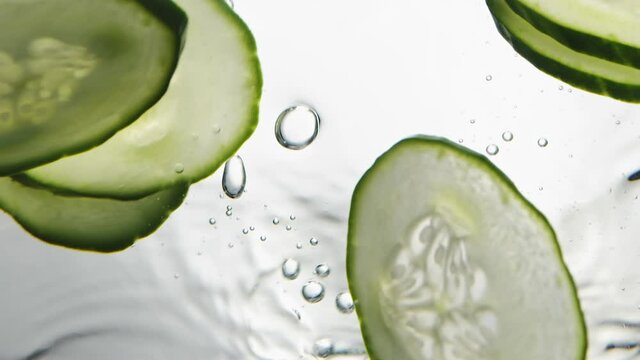 Thin slices of fresh cucumber fall down into clear water with small bubbles on white background extreme close bottom view slow motion