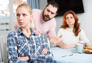 Upset girl sitting at table after quarrel and man with mother on background at home