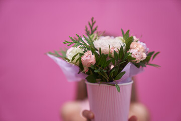 A little girl in pink dress with bouquet of mixed flowers in a paper cup, hid behind the flowers. The girl holds out her hands and gives flowers.