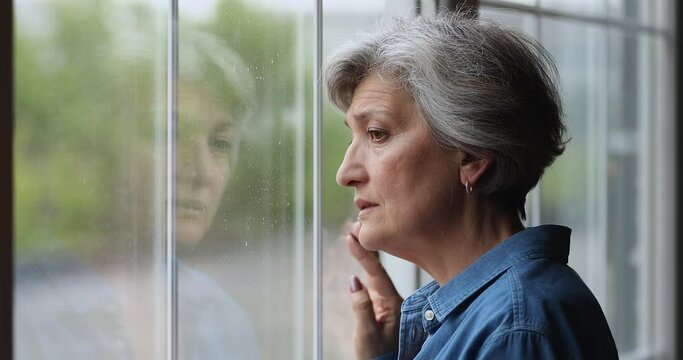 Head shot frustrated unhappy unhealthy middle aged hoary retired woman looking out of window, suffering from negative melancholic thoughts or feeling lonely alone indoors, ageing process concept.
