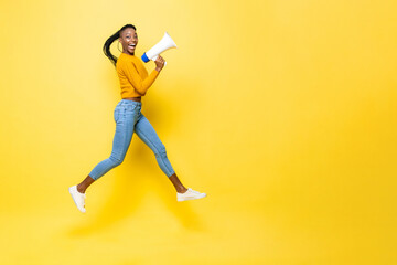 Fototapeta na wymiar Jumping portrait of ecstatic smiling young African American woman with megaphone in isolated studio yellow background with copy space