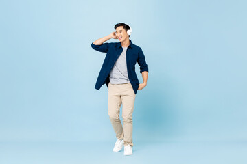 Full body portrait of smiling young handsome Asian man listening to music with wireless headphones...