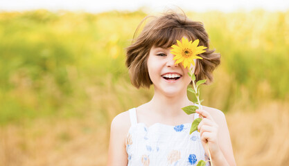 Cute child girl wear dress with sunflower in summer field. Happy little girl hide eye with sunflower. Summer time concept.