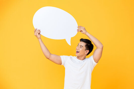 Portrait of cheerful smiling young handsome Asian man holding and looking up at speech bubble with empty space for text on colorful yellow studio background