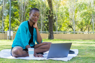 black african woman sitting on a blanket in the park using the computer, with her cup of coffee and headphones next to her.