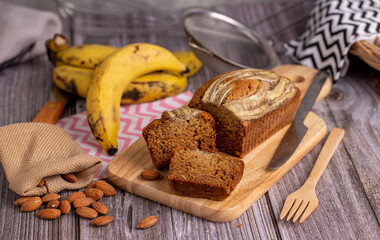 Homemade banana bread sliced on a wooden board with almond, banana and cooking tool on table.