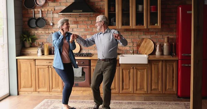 Full length overjoyed older senior couple dancing to energetic music in old-fashioned kitchen, celebrating special event, marriage anniversary or enjoying carefree active weekend together at home.
