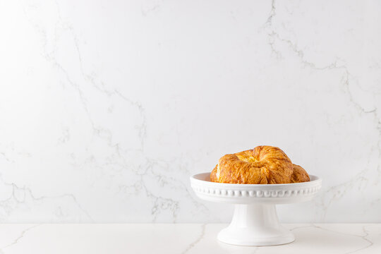 A few croissants in a white cake stand placed on a white marble looking quartz countertop, against a white marble background