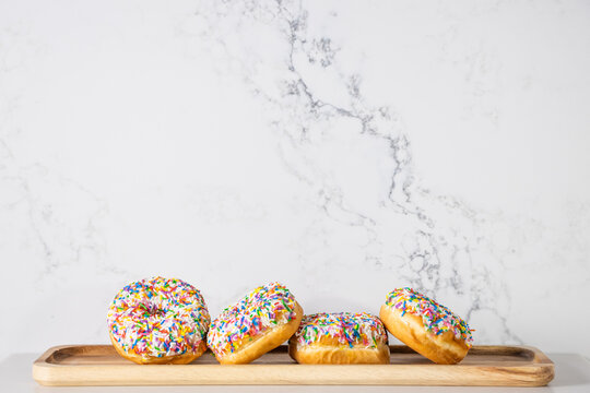 A few donuts covered with colourful sprinkles laying in a wooden tray against a marble looking background made of quartz