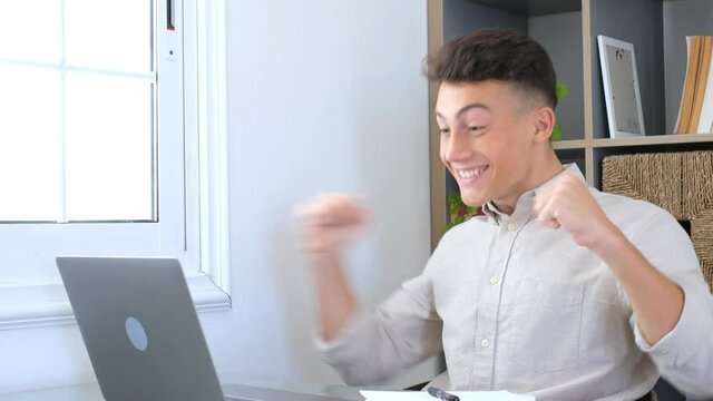 Overjoyed caucasian ethnicity teenager sit at desk looks at laptop screen read incredible news clench fists makes yes gesture celebrate on-line lottery gambling win, getting new job offer feels happy
