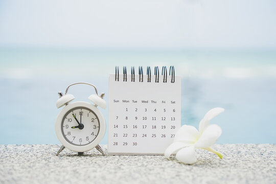 white calendar and alarm clock on concrete floor with blurred plumeria flower and seascape background for recreation or work from home concept
