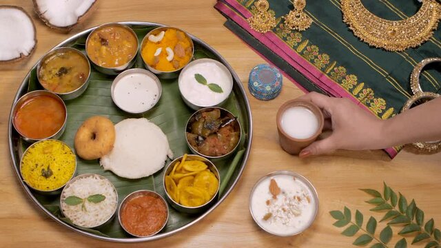 Female hands keeping a glass of buttermilk (Chaas) near a South Indian Thali. A plate full of traditional food like Vada  Idli  lemon rice  banana chips  Sambhar served on a table with jewelry and ...