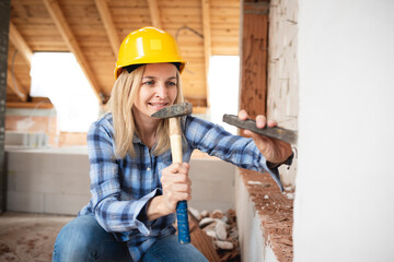 pretty young worker woman with blue work shirt and yellow protective helmet works on construction site and holds hammer in her hand, concept of female worker