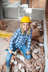 pretty young worker woman with blue work shirt and yellow protective helmet works on construction site and holds hammer in her hand, concept of female worker