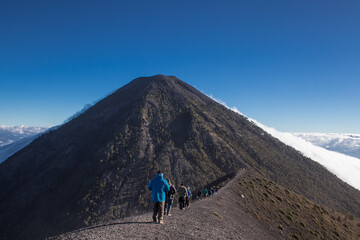 general shot of a group of hikers walking in a row on the trail with a view of the Acatenango Volcano and the camping area from the ridge of the Fuego Volcano on a beautiful sunny blue sky morning