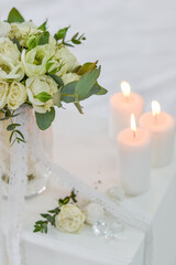 A wedding bouquet of white roses and eucalyptus stands in a vase on the table next to candles and sweets. The bride's morning preparations. Details of a stylish European wedding day