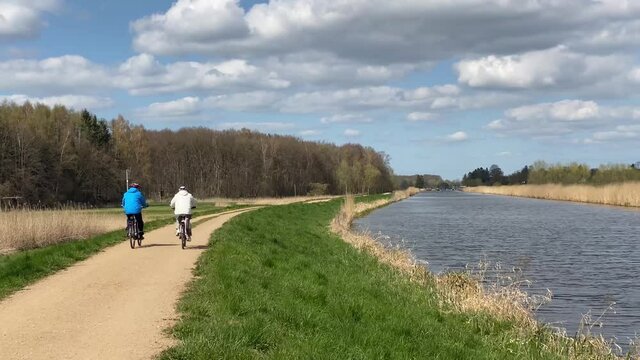 Aktiv im Alter. Zwei ältere Menschen, Senioren fahren mit dem Fahrrad an einem sonnigen, schönen Tag an einem Kanal, Fluss, Gewässer entlang.