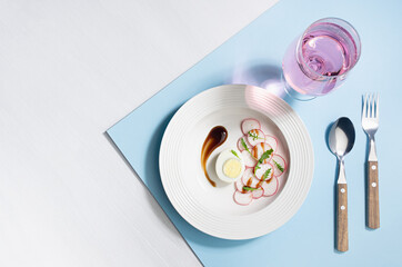Colorful organic salad of radish, arugula, eggs with balsamic sauce with rose sparkling wine in glass on blue background, top view, border. Modern concept with shadow and hard light.